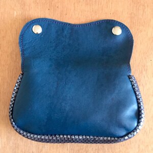 May include: A blue leather pouch with a decorative beaded trim and two silver snap closures. The pouch is open, revealing the interior. The stitching is a contrasting purple color. The pouch is on a wooden surface.