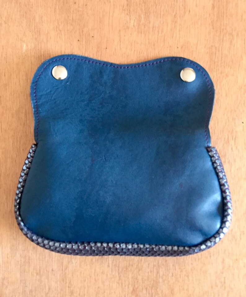 May include: A blue leather pouch with a decorative beaded trim and two silver snap closures. The pouch is open, revealing the interior. The stitching is a contrasting purple color. The pouch is on a wooden surface.