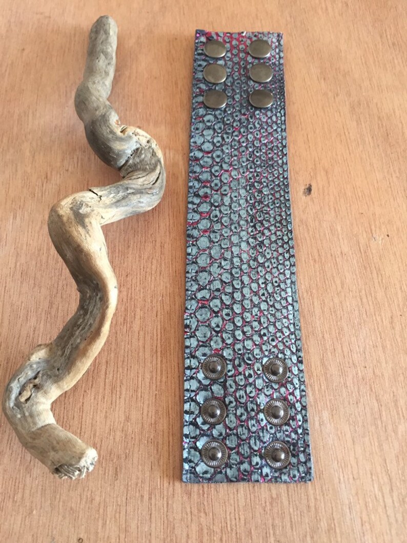 Op de afbeelding: Een brede, getextureerde armband met een teal en rood slangenpatroon en zes metalen drukknoopsluitingen. Een stuk drijfhout ligt naast de armband op een houten oppervlak. De armband is ongeveer 5 cm breed.