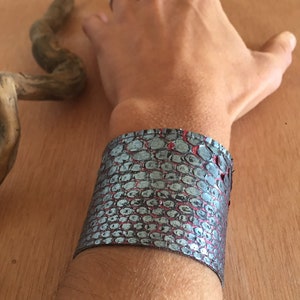 Op de afbeelding: Een brede manchetarmband met een textuurpatroon in grijstinten en rood. De armband is om een pols gewikkeld en toont het ontwerp. De manchet is een opvallend accessoire, geschikt voor verschillende stijlen.