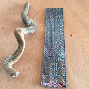 Op de afbeelding: Een leren manchetarmband met een slangenpatroon in teal, roze en zwart, vastgezet met zes bronskleurige drukknopen. Een stuk drijfhout ligt naast de armband op een houten oppervlak.
