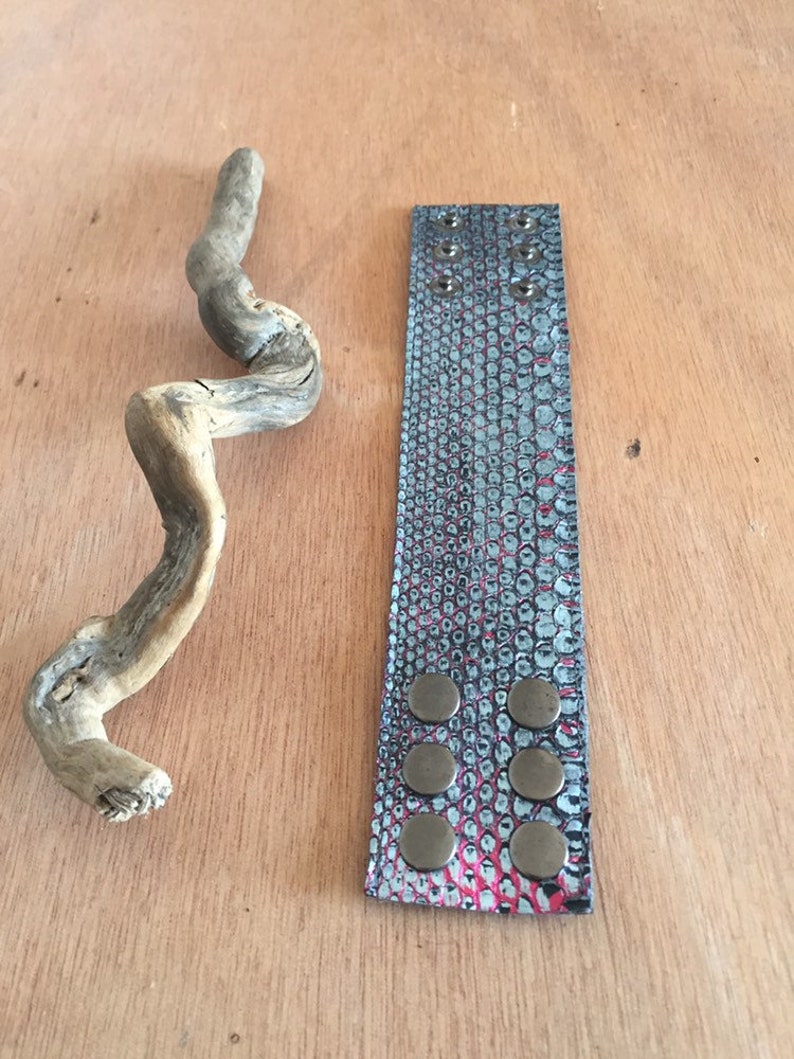Op de afbeelding: Een leren manchetarmband met een slangenpatroon in teal, roze en zwart, vastgezet met zes bronskleurige drukknopen. Een stuk drijfhout ligt naast de armband op een houten oppervlak.