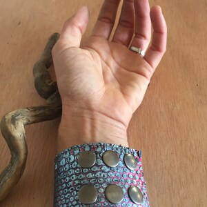 Op de afbeelding: Een brede manchetarmband met een slangenleerpatroon in tinten blauw en roze. De armband is versierd met zes grote, ronde, zilverkleurige studs. De manchet wordt om een pols gedragen.