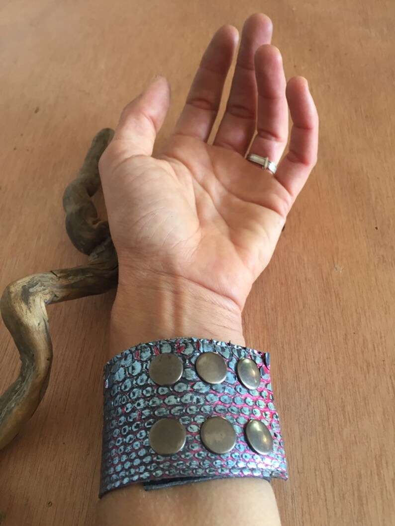 Op de afbeelding: Een brede manchetarmband met een slangenleerpatroon in tinten blauw en roze. De armband is versierd met zes grote, ronde, zilverkleurige studs. De manchet wordt om een pols gedragen.