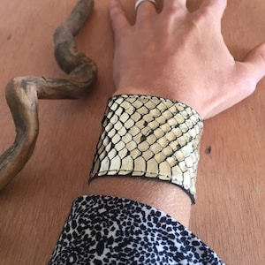 Peut inclure: Un large bracelet manchette en matériau à motif peau de serpent crème et noir. Le bracelet est porté sur un poignet, avec une manche à motifs noirs et blancs visible en dessous. Un morceau de bois flotté est en arrière-plan.