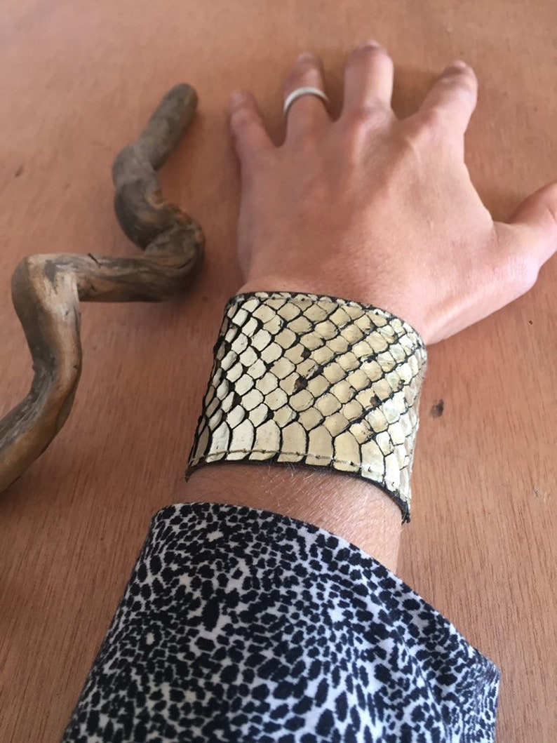 Op de afbeelding: Een brede manchetarmband van cr&egrave;me- en zwart slangenleer-patroonmateriaal. De armband wordt om een pols gedragen, met een zwart-wit patroonmouw eronder zichtbaar. Een stuk drijfhout staat op de achtergrond.