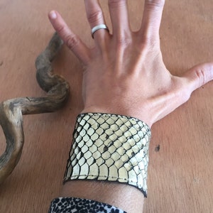 Op de afbeelding: Een brede manchetarmband van cr&egrave;me en zwart slangenleer. De armband wordt om de pols gedragen en het slangenleerpatroon is zichtbaar. Een opvallend accessoire.