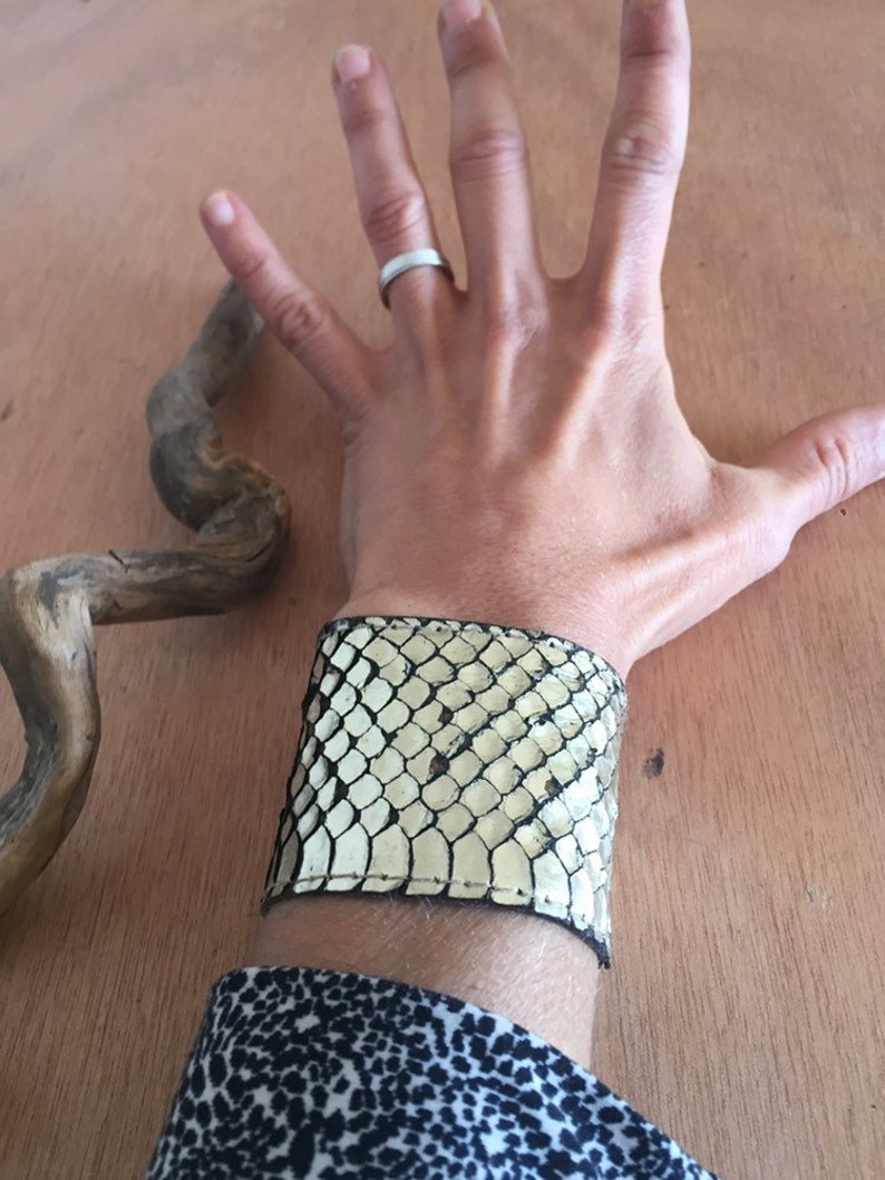 Op de afbeelding: Een brede manchetarmband van cr&egrave;me en zwart slangenleer. De armband wordt om de pols gedragen en het slangenleerpatroon is zichtbaar. Een opvallend accessoire.