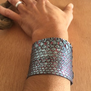 Op de afbeelding: Een brede manchetarmband met een slangenpatroon in grijstinten en rood. De armband wordt om de pols gedragen en toont het gestructureerde oppervlak en het ontwerp. De armband lijkt van leer of een vergelijkbaar materiaal te zijn.