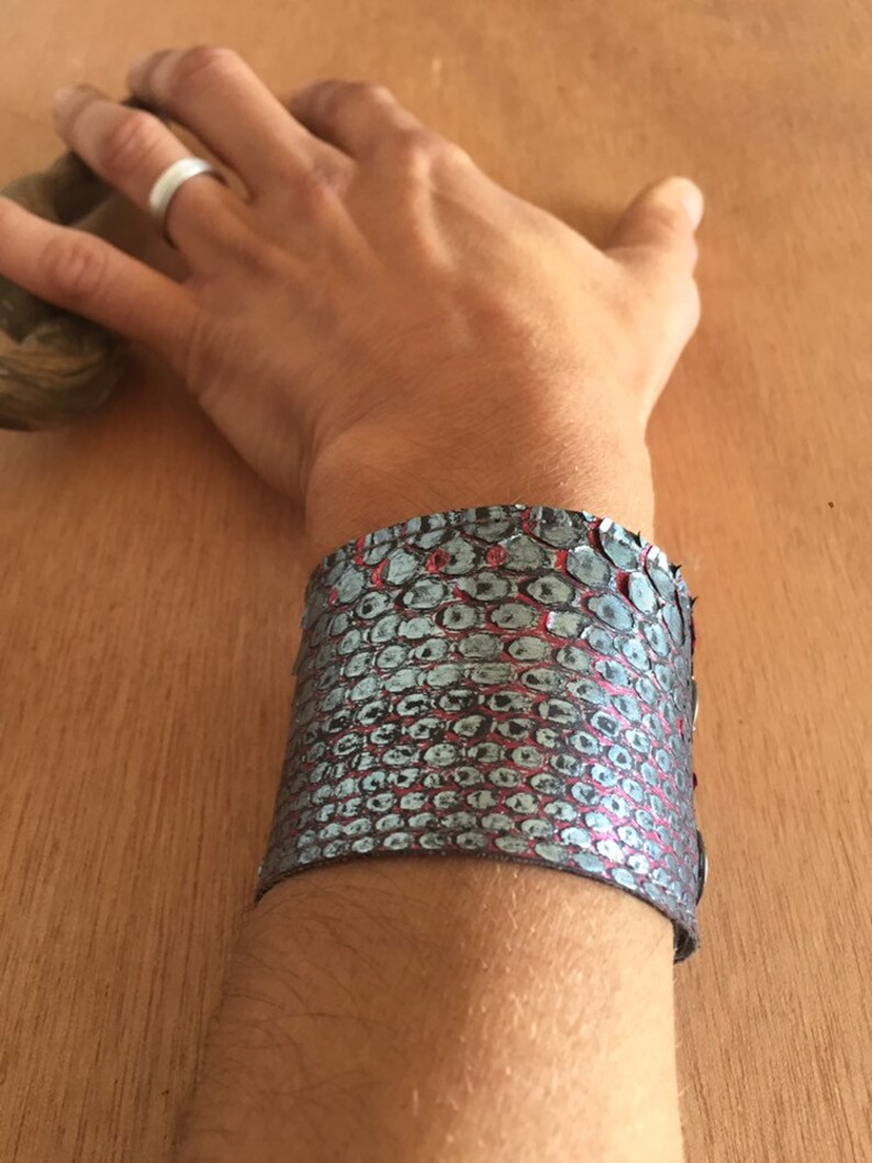 Op de afbeelding: Een brede manchetarmband met een slangenpatroon in grijstinten en rood. De armband wordt om de pols gedragen en toont het gestructureerde oppervlak en het ontwerp. De armband lijkt van leer of een vergelijkbaar materiaal te zijn.