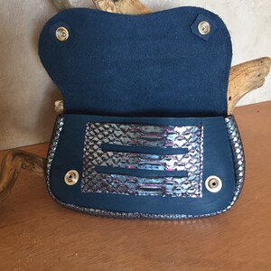 May include: Open navy blue clutch with a silver and purple snakeskin-patterned accent. The clutch has a flap closure with snap fasteners and a suede interior. The bag is displayed on a wooden surface.