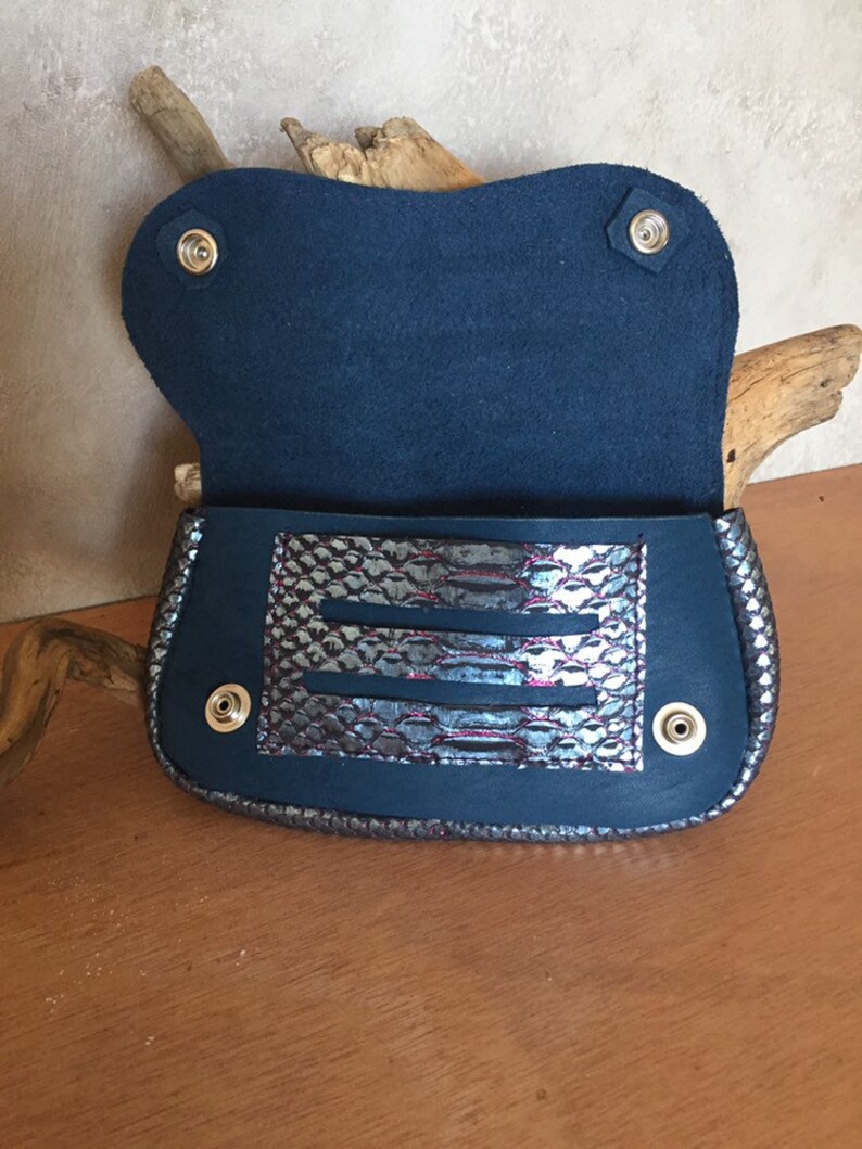 May include: Open navy blue clutch with a silver and purple snakeskin-patterned accent. The clutch has a flap closure with snap fasteners and a suede interior. The bag is displayed on a wooden surface.