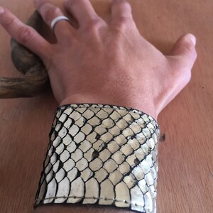 Op de afbeelding: Een brede manchetarmband van cr&egrave;me en zwart slangenleer. De armband is om een pols gewikkeld en toont de textuur en het patroon van het slangenleer. De manchet is een opvallend accessoire.