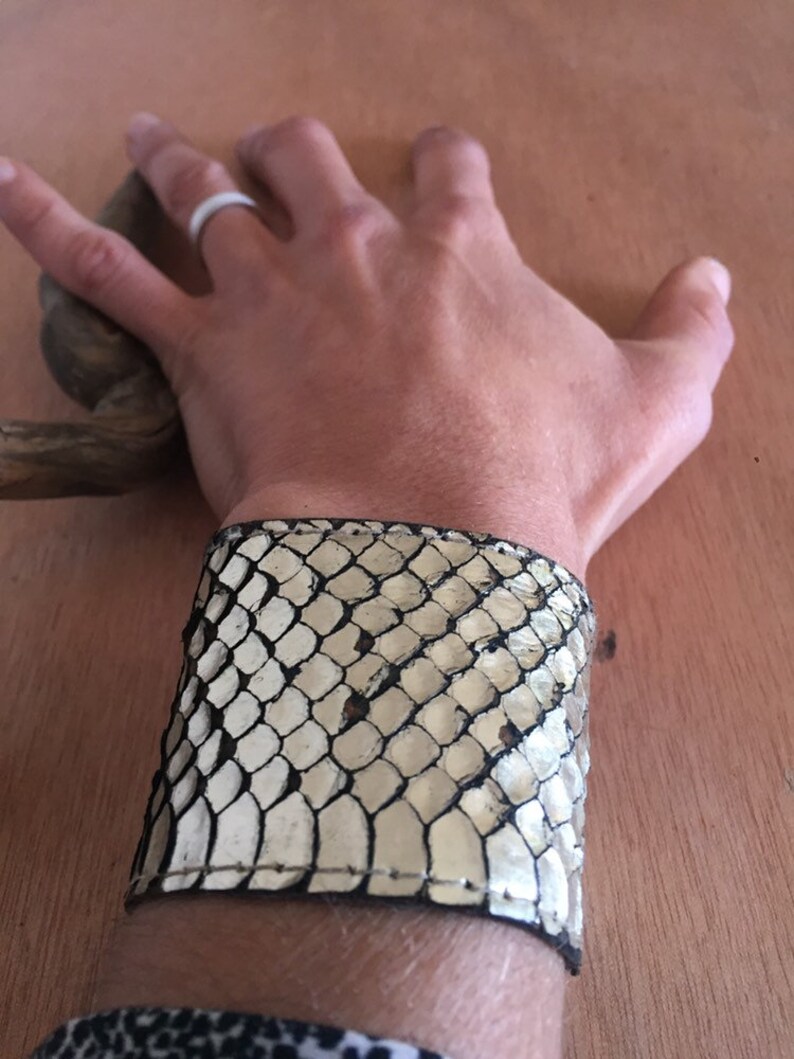 Op de afbeelding: Een brede manchetarmband van cr&egrave;me en zwart slangenleer. De armband is om een pols gewikkeld en toont de textuur en het patroon van het slangenleer. De manchet is een opvallend accessoire.