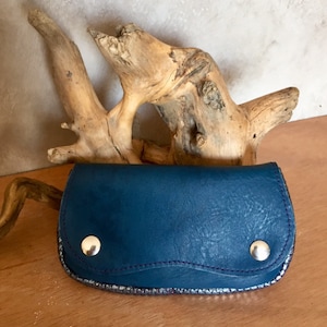 Peut inclure: Un portefeuille en cuir bleu sarcelle avec une bordure argentée et deux fermetures à pression argentées. Le portefeuille est posé sur une surface en bois avec un morceau de bois flotté en arrière-plan. La couture est violette.