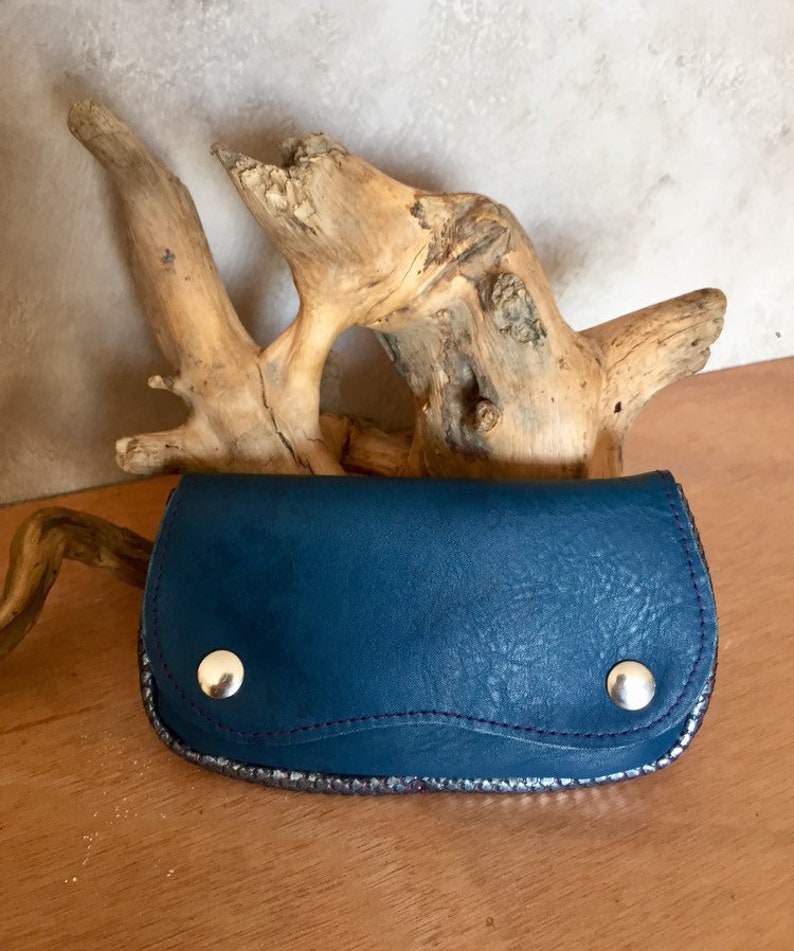 May include: A teal leather wallet with silver trim and two silver snap closures. The wallet is on a wooden surface with a piece of driftwood in the background. The stitching is purple.