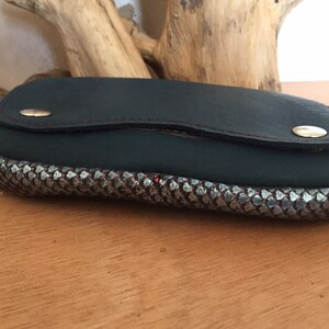 May include: A dark green leather eyeglass case with a silver and black patterned border. The case has a flap closure with a gold-colored snap. Red stitching accents the leather.
