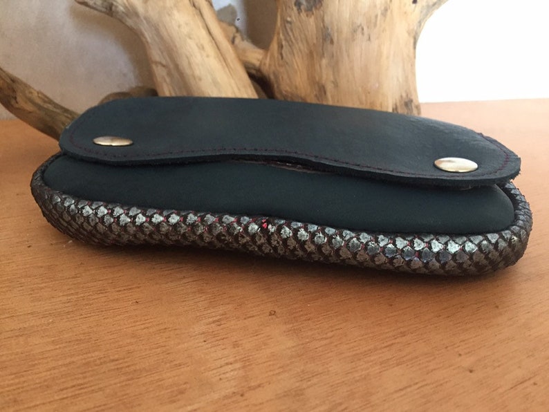 May include: A dark green leather eyeglass case with a silver and black patterned border. The case has a flap closure with a gold-colored snap. Red stitching accents the leather.