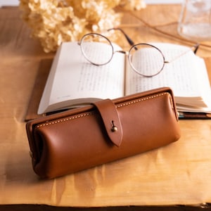 以下が含まれることがあります： 木製のテーブルの上に置かれた、茶色の革製のペンケース。フラップ式で真鍮製のボタンが付いています。ケースの隣には本と眼鏡があります。