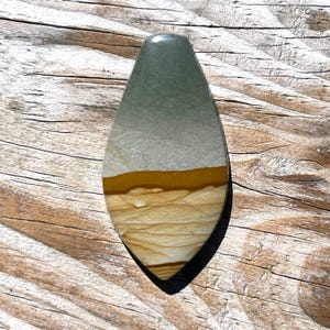 Könnte beinhalten: Ein tropfenförmiger, polierter Stein. Die Oberseite ist ein gedämpftes Grün, das in Grau übergeht, dann ein goldenes Band und schließlich ein geschichtetes beige-braunes Muster. Der Stein liegt auf einem Holzgrund.
