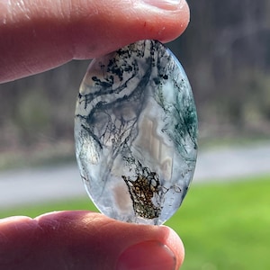May include: An oval moss agate gemstone with a translucent base. The stone features intricate, tree-like inclusions in shades of green, black, and brown, creating a natural, artistic pattern. The polished surface reflects light, highlighting the internal details.