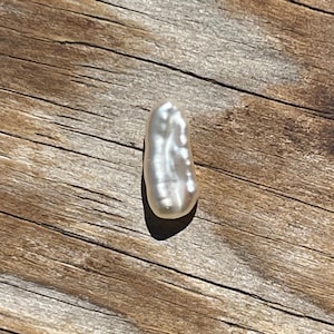 May include: A lustrous, off-white baroque pearl with a unique, elongated shape. The pearl reflects light, showcasing a subtle iridescence. The pearl is set against a weathered, wooden surface, highlighting its natural beauty.