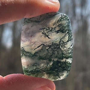 May include: A rectangular moss agate gemstone with rounded corners. The stone has a translucent base with green, black, and white inclusions, resembling a landscape. The stone is held between fingers.