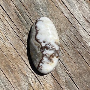 May include: Oval-shaped polished stone with a white base and brown and gray veining. The stone is smooth and has a glossy finish, resting on a weathered wooden surface.