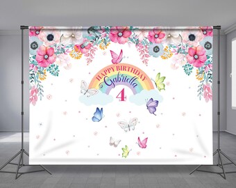 May include: A white backdrop with a rainbow and clouds, featuring the text "Happy Birthday Gabriella 4". The backdrop is decorated with pink, blue, and yellow flowers and butterflies.