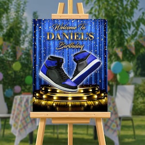May include: A blue and black birthday party sign with a blue curtain backdrop and gold confetti. The sign says "Welcome to Daniel's Birthday" and features two blue and black sneakers on a stage with gold lights.
