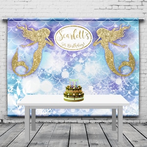 May include: A mermaid-themed birthday party backdrop with a white table and a cake. The backdrop features two gold glitter mermaids, a blue and purple scale pattern, and the text "Scarlett's 5th Birthday!". A two-tiered cake with candles sits on the table.