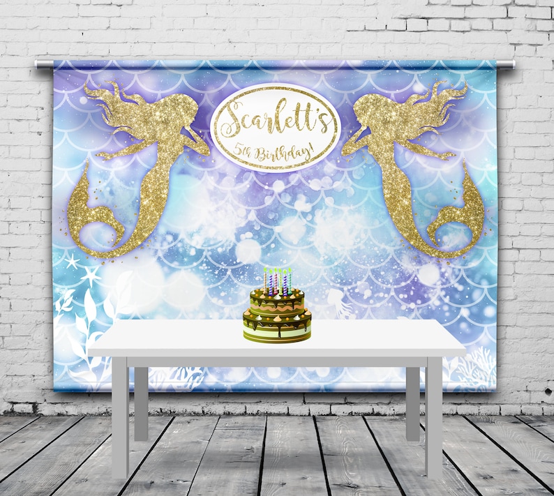May include: A mermaid-themed birthday party backdrop with a white table and a cake. The backdrop features two gold glitter mermaids, a blue and purple scale pattern, and the text "Scarlett's 5th Birthday!". A two-tiered cake with candles sits on the table.