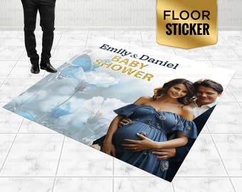 May include: A blue and white floral baby shower floor sticker with the text "Emily & Daniel Baby Shower" in gold glitter lettering. The sticker features a couple, the woman is pregnant and wearing a blue dress, and the man is wearing a white shirt and black pants.