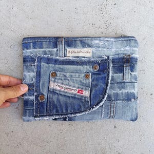 Può includere: Una pochette rettangolare in denim realizzata con jeans blu riciclati, dotata di una tasca frontale con etichetta Diesel Industry, rivetti color rame, bordi sfrangiati e una piccola etichetta in tessuto con la scritta Il filo del riciclo.