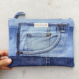 Può includere: Una pochette rettangolare in denim realizzata con jeans blu riciclati, caratterizzata da una tasca frontale con cuciture a vista, rivetti in metallo e un bordo sfrangiato. Una piccola etichetta in tessuto bianco con la scritta "il filo del riciclo" è cucita sul davanti e una chiusura a zip si trova sulla parte superiore.