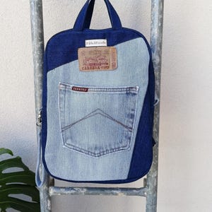 Puede incluir: Mochila vaquera con diseño patchwork. La bolsa presenta un bolsillo de mezclilla azul claro y un cuerpo de mezclilla azul oscuro. Un parche de cuero con el texto "CARRERA JEANS" está cosido en la parte delantera.