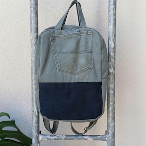 Può includere: Uno zaino in denim con una sezione superiore grigio-verde sbiadito e una sezione inferiore blu navy scuro. Lo zaino ha un manico, spallacci regolabili e una tasca frontale. La borsa è appesa a una scala di metallo.