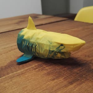 以下が含まれることがあります： 黄色と青色の3Dプリントされたサメの置物。サメは幾何学的なデザインでスタイリッシュに作られており、大理石のような効果があります。