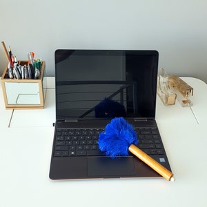 Może przedstawiać: Czarny laptop z niebieskim pędzlem do kurzu na klawiaturze. Laptop jest otwarty, a ekran jest czarny. Laptop stoi na białym biurku.