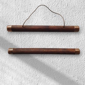 Op de afbeelding: Twee houten posterhangers met koperkleurige eindkappen en een dun bruin touwtje om op te hangen. Het hout heeft een rijke, donkerbruine kleur. De hangers zijn ontworpen om kunstwerken of posters weer te geven.