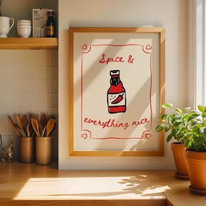 Puede incluir: Un estampado enmarcado con las palabras "Spice & everything nice" en escritura roja. El estampado presenta una ilustración de dibujos animados de una botella de salsa picante roja con un chile. El marco es de color marrón claro.