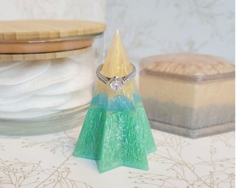 May include: A green, blue, and yellow resin ring holder shaped like a star. A silver ring with a clear stone is on the holder.
