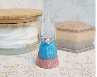 May include: A blue and pink ring holder shaped like a cone with a silver ring with a clear stone on top. The ring holder is sitting on a white surface with a floral pattern.