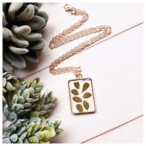 May include: A gold chain necklace with a rectangular pendant featuring pressed green leaves encased in clear resin.