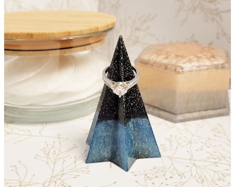 May include: A black and blue glitter ring holder shaped like a pyramid. The ring holder is made of resin and has a sparkly finish. A silver ring with a clear stone is displayed on the holder.