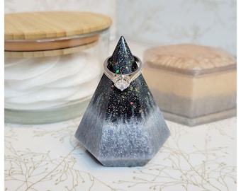 May include: A geometric ring holder in shades of black and gray, with a sparkling finish. A silver ring with a large, clear stone sits atop the holder. The holder is a decorative item for jewelry.