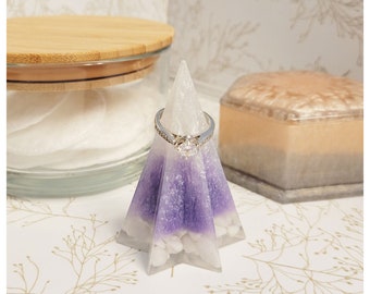 May include: A white and purple ring holder shaped like a pyramid with a silver ring on top. The ring holder is made of resin and has a sparkly finish.