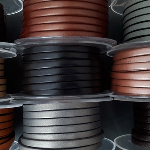 Könnte beinhalten: Spulen mit flachem Lederband in verschiedenen Farben, darunter Braun, Schwarz, Beige und Grau. Das Band ist ordentlich auf transparenten Kunststoffspulen aufgewickelt. Ideal für Schmuckherstellung und Bastelarbeiten.