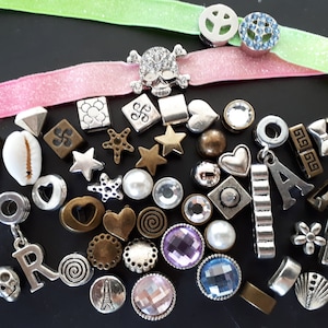 Op de afbeelding: Een verzameling diverse metalen en kristallen bedels, kralen en tussenstukjes in verschillende vormen zoals sterren, harten, schedels en letters, samen met twee fluwelen linten in lichtgroen en roze met vredesteken- en schedelversieringen.