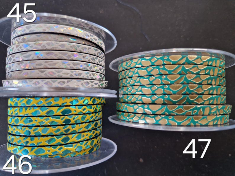 May include: Three spools of flat leather cord featuring textured patterns. Spool 45 has a white base with iridescent scales. Spool 46 is yellow with teal scale-like shapes. Spool 47 is teal with gold-colored metallic scale-like shapes.